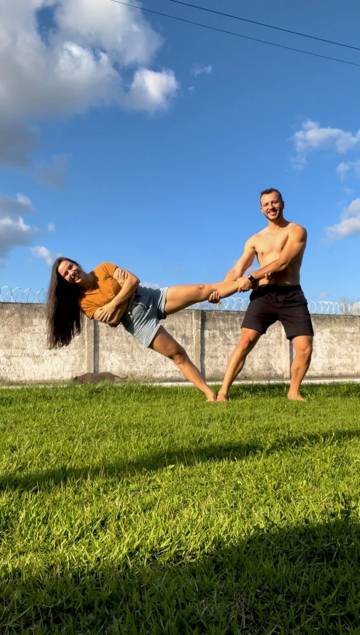Roberta e Eric fazendo uma acrobacia num gramado, sob céu azul