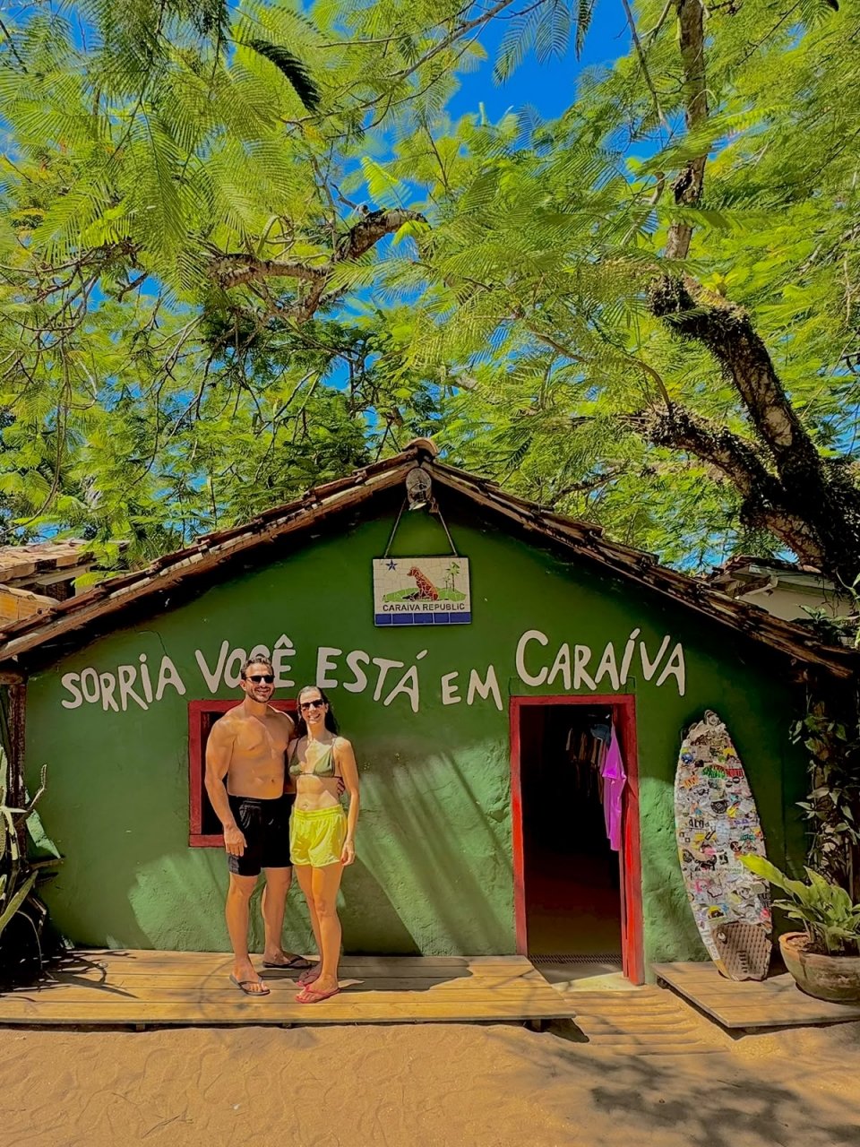 Roberta e Eric em frente a uma casinha verde com a frase Sorria, você está em Caraíva