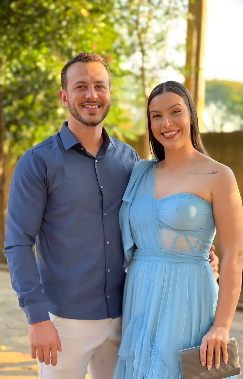 Eric de camisa azul e Roberta de vestido azul-claro, lado a lado, sorrindo ao ar livre