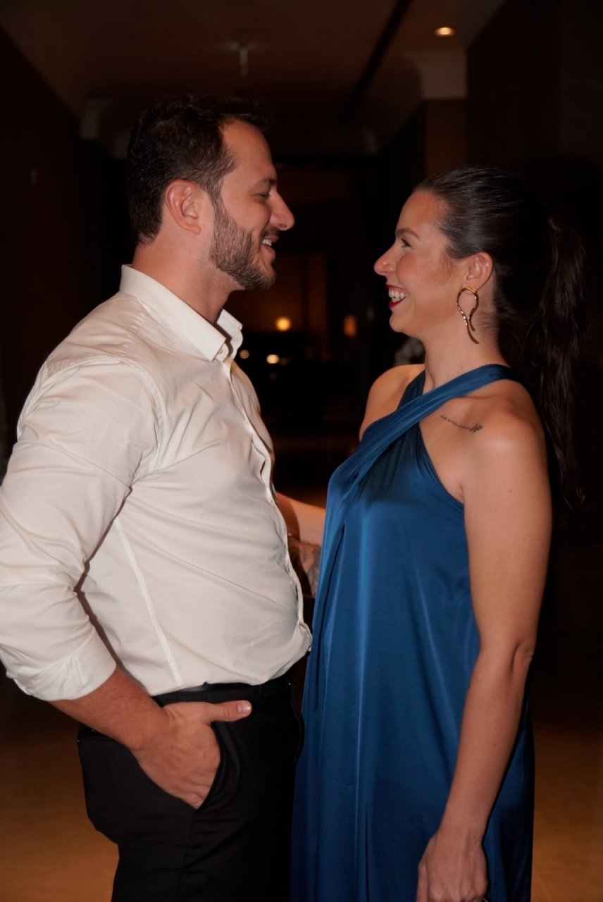 Roberta e Eric frente a frente, sorrindo um para o outro, em traje de festa num ambiente à noite