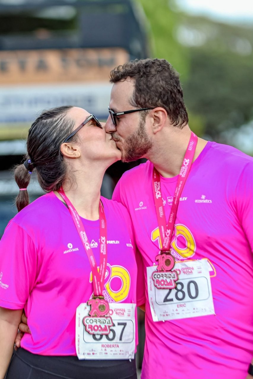 Roberta e Eric com camisetas rosa e medalhas de corrida, trocando um beijo