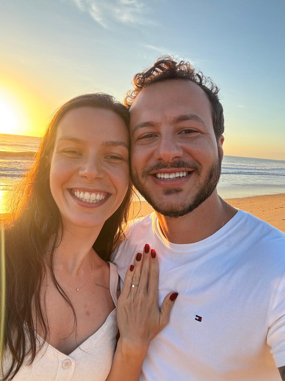 Roberta e Eric sorrindo na praia ao entardecer, ela mostrando a aliança de noivado