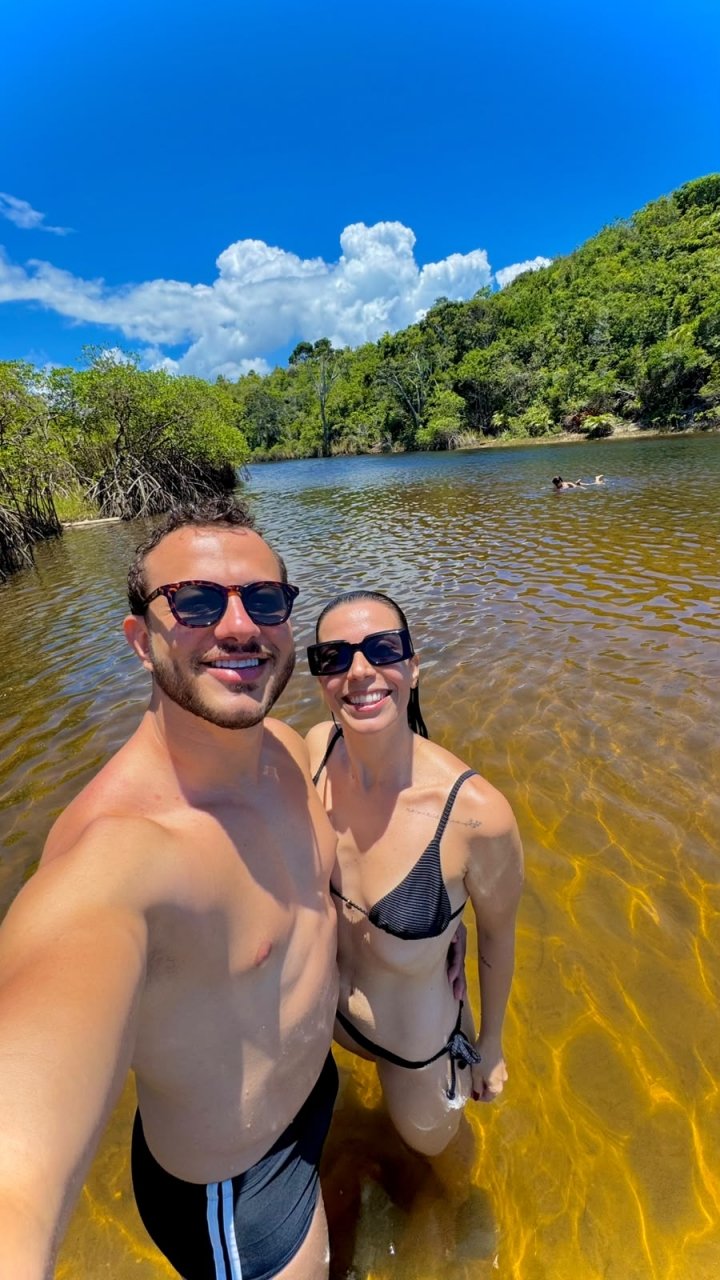 Selfie de Eric e Roberta, de óculos escuros, dentro de um rio de água clara cercado de mata